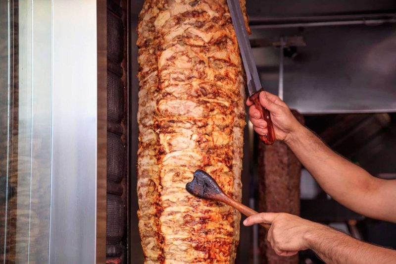 Gyros or doner, Middle East traditional food. Man's hands holding a knife, cutting slices from a vertical skewer